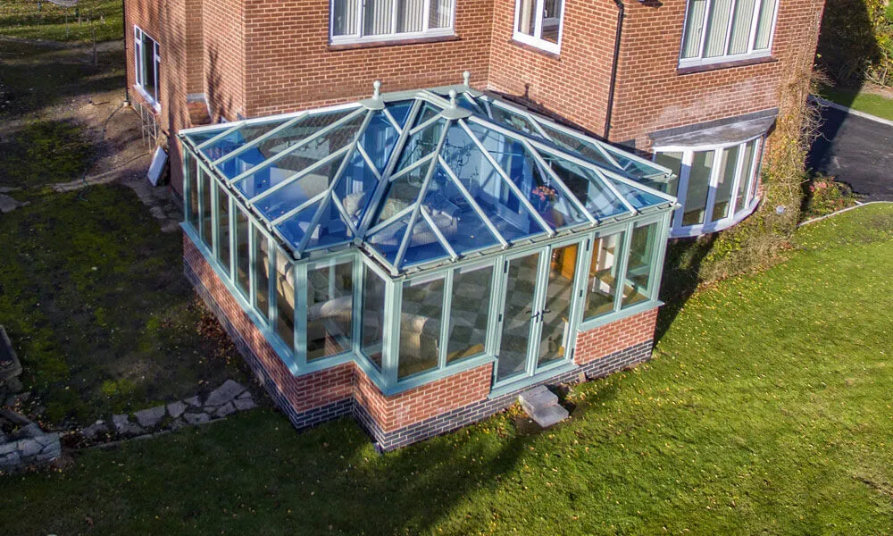 High-angle aerial drone view of a bespoke, Chartwell green uPVC conservatory built in an Edwardian style, featuring a multi-faceted glass roof and double doors, attached to a red brick house with a green lawn.