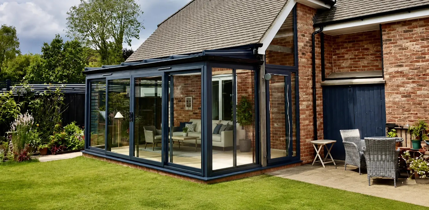 Combination conservatory {location} with black-framed glazing, sliding doors, patio furniture and garden views attached to a red brick house.