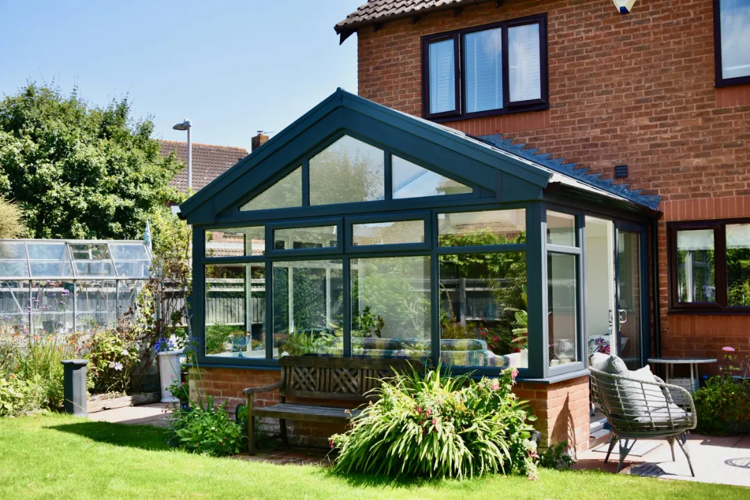 Dark green gable end conservatory attached to a brick house, with garden planting and a bench in front.