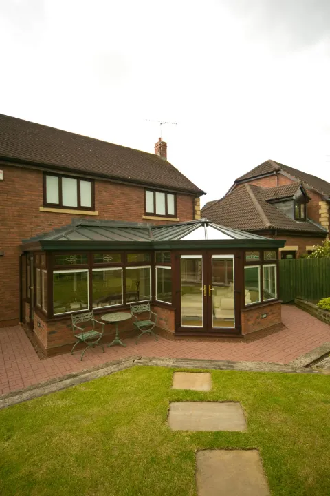 Large combination conservatory with brown frames, brick base, glazed roof and double doors facing a garden.